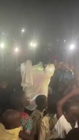 The video shows a performer in a shiny white outfit and a head covering singing into a microphone while surrounded by a crowd of people, including many children. The lighting is bright, with what appear to be spotlights shining towards the camera. People are clapping, cheering, and reaching out towards the performer. The scene appears to be taking place outdoors at night.