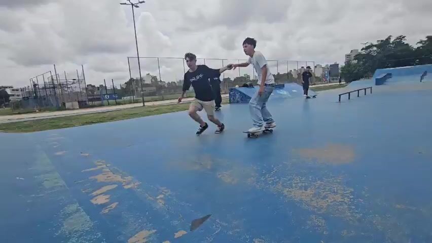 Two young men are at a skate park. The skate park has multiple ramps. The first man wearing a white T-shirt and loose-fitting jeans pushes himself on a skateboard to a ramp. The second man wearing a black T-shirt and shorts runs to catch up. At the ramp, the second man helps the first man to skateboard up the ramp. The first man, now at the top of the ramp, grabs the second man's hand as the second man climbs to the top. Once at the top of the ramp together, the first man skateboards down the ramp back to the flat skate park surface. The second man runs down the ramp with him. There are other people at the skate park, including one person on a skateboard and a dog in the background. The sky is overcast.