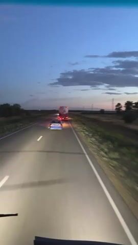 This media file captures a view from a vehicle traveling on a highway at dusk. The primary subjects are a white sedan and a large truck with a trailer, both illuminated by their headlights and taillights, respectively. The sedan is in the left lane, and the truck is further ahead in the right lane. The highway stretches into the distance, bordered by dry, grassy terrain and scattered trees. Utility poles line the side of the road.

The activity is vehicular travel on a highway. The time of day is twilight, as indicated by the deep blue sky with wispy clouds tinged with orange and pink near the horizon. The lighting suggests a fading sunset or the approaching night. The weather appears clear, with no rain or fog visible.

Notable details include the bright headlights of the vehicle from which the footage is filmed, casting a strong glow on the road ahead and creating tire tracks on the asphalt. The truck's taillights are red, and the sedan has blue accents, possibly from its lights. The landscape is sparse and rural. The mood conveyed is one of quiet travel, perhaps a long journey as night falls. There is no visible text in the media file. The location context provided is Baiqadam, Kazakhstan.