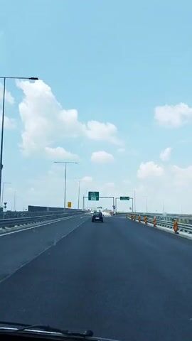 The video captures a point-of-view perspective from a vehicle driving on a multi-lane highway under a bright blue sky with scattered white clouds. The road is paved and appears to be in good condition, with clear white lane markings. In the distance, there are several large green highway signs mounted on gantries that arch over the road. These signs display directional information, indicating exits and destinations. One sign shows "Ploiesti", "Urziceni", "Bucuresti", and "Centura Titan". Another sign to the right indicates "Constanta". There are also overhead light poles and barriers along the sides of the highway. A few other vehicles, including a dark-colored car and a white truck, are visible on the road, some ahead of the viewer's vehicle and some in adjacent lanes. The overall environment suggests a modern highway infrastructure in a generally clear and open landscape.