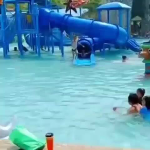 The video shows a water park with a large blue play structure featuring slides and a tunnel that empties into a shallow pool. Several children are swimming in the pool and playing around the slide. A child can be seen exiting the tunnel of the slide. Other park visitors are visible in the background. There are various inflatable toys and water guns near the pool edge in the foreground. The water in the pool appears clean and blue. The video is taken from a slightly elevated position near the pool's edge.