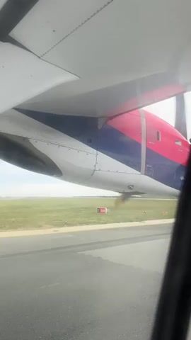 This is a first-person perspective video taken from inside an airplane, likely during taxiing or takeoff. The view is dominated by the underside of the wing and a portion of the aircraft's fuselage, painted with bold red, white, and blue stripes. A propeller, blurred by its rotation, is visible in the foreground.

Outside the window, a runway and a grassy field are visible under a cloudy sky. A runway marker with the designation "30R" is clearly visible on the field, indicating a specific runway direction. The overall lighting suggests it is daytime. The scene is set at an airport, likely in Surcin, Serbia, given the provided location context. There are no people visible in the frame. The image conveys a sense of anticipation or the beginning of a journey.