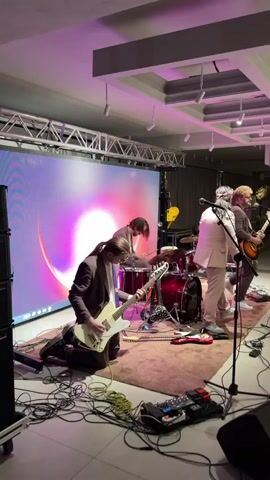 A band performs on a stage with a large LED screen displaying a pink and purple abstract graphic. The band members include a bassist in the foreground playing a white bass guitar while kneeling, a drummer in the middle playing a red drum set, and two guitarists on the right. The guitarist in the middle is wearing a light-colored suit and the guitarist on the far right is wearing a dark shirt and appears to be singing. The stage is lit with pink and purple lights. Cables and musical equipment are visible on the floor in front of the stage. The overall mood appears energetic and focused on the performance. There is no visible text in the media file. The location is specified as Kaji-Sai 2, Kyrgyzstan. The time of day is not specified, but the lighting suggests it is indoors and likely evening or night, consistent with a performance setting.