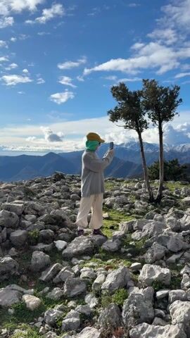 The video shows a person taking pictures with a smartphone of a mountain range. The person is wearing a light-grey sweater, light-colored pants, a green head covering, and a baseball cap. The mountain range is in the background and has snow-capped peaks. There are some small trees on the rocky ground where the person is standing. The sky is blue with some white clouds.