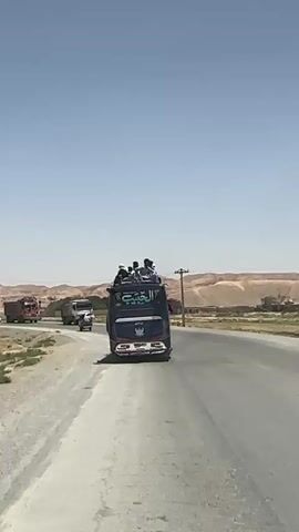 A blue bus with several people riding on its roof drives on a paved road under a clear, bright blue sky. The bus is in the foreground, viewed from behind, and has Arabic or Urdu script on its rear, possibly reading "الغنّاء". To the left, a red/brown truck is visible. As the video progresses, a white truck with "EK-BUILDERS" written on its side appears on the right and overtakes the blue bus. Another white car and a motorbike are also seen further back on the road. The setting appears to be a dry, arid region with light-colored hills or mountains in the distant background. Utility poles line the side of the road. The time of day is sunny, with strong light indicating midday. The road itself appears a bit dusty on the edges. The vehicles are moving on what seems to be a main road connecting different areas.