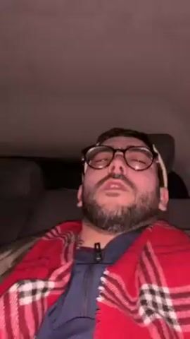 A man with a beard and round glasses is lying down inside a car. He is wearing a blue jacket and a red plaid scarf. His eyes are closed at first, but then he opens them and looks up. He moves his hand slightly. The interior of the car is visible. The overhead is grey.  The headrest of a car seat is visible behind him.