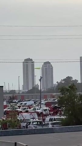 This is a video of a parked lot filled with many vehicles, most of which appear to be white vans and trucks. In the background, there are two tall, rectangular buildings with white facades and dark vertical accents. There is a green flag or streamer attached to a pole in front of the buildings, which is blowing in the wind. Overhead, there are power lines. The sky is overcast.