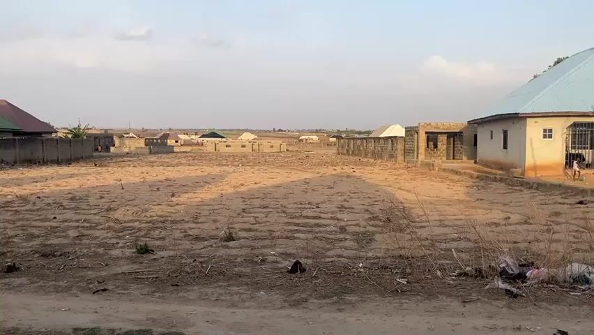 The video depicts a scene of a sparsely populated area with several buildings in various stages of construction. The foreground is dominated by a large, barren plot of land covered with dry soil, sparse vegetation, and scattered debris. In the background, there are a few houses, some of which appear to be inhabited, while others seem to be unfinished structures. A concrete block wall surrounds some of the properties, and the buildings are a mix of completed and partially constructed forms, with visible brickwork and corrugated roofing. The sky is light blue, indicating it is daytime, with a few wispy clouds. The general atmosphere suggests a developing or rural area. A child appears in the right corner of the image.