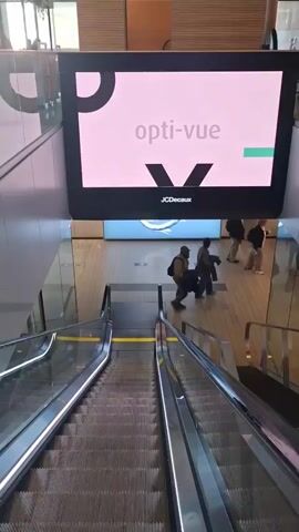 The video shows an escalator moving downwards, with people walking around in a well-lit area. An advertising screen above the escalator displays an advertisement for "opti-vue" and shows the locations of 11 shops. The ad cycles through different images, including a woman wearing glasses and text listing the locations. Below the screen is another advertisement with a blue background.