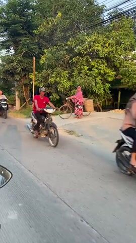 A video shows a street scene. In the foreground, a person's hand and handlebars of a motorcycle are visible, indicating the footage is from the perspective of someone riding a motorcycle. The motorcycle is moving along a paved road. On the left side of the road, there is a grassy area with trees and some buildings. A woman is riding a black scooter towards the viewer. Further down the road, a woman in a pink hijab and a floral skirt is standing next to a bicycle, also facing the viewer. On the right side of the road, another motorcycle is moving away from the viewer. Beyond the immediate roadside, there are more trees, buildings, and power lines crisscrossing the sky. Several flags, some red and white, are planted near a building on the left side of the road. The lighting suggests it is daytime, likely late afternoon given the warm tones.
