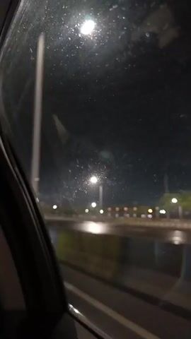 The video shows a nighttime view from inside a car, looking out the passenger window. The window is dirty and streaked, with a few bright lights reflecting on its surface. Outside, the scene is dark with several streetlights illuminating a road and some trees. There are yellow and black barriers along the side of the road. The car appears to be moving, as suggested by the slight blur of the lights and the perspective shifting. The overall atmosphere is one of a quiet, possibly lonely, night drive.