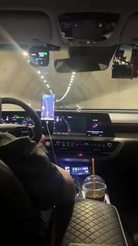 This media file shows the interior of a car driving through a tunnel. The camera is positioned in the front passenger seat, looking forward at the dashboard and the tunnel ahead. The driver's hands are visible on the steering wheel, and they are wearing a dark-colored shirt.

On the dashboard, there are several electronic devices. A smartphone is mounted on a stand, displaying a navigation app with a route highlighted. To the left of the phone, a digital display shows "98." To the right of the phone, the car's infotainment screen shows a navigation map. Below the infotainment screen, there are climate control buttons and a small digital display showing "21,080." A cup of iced coffee with a straw sits in the center console.

The tunnel appears to be well-lit with overhead lights. The walls of the tunnel are textured, suggesting it might be an older or naturalistic tunnel construction. The overall lighting suggests it is daytime, but the enclosed space of the tunnel makes it dark.

The scene is focused on the act of driving through the tunnel. The presence of navigation devices and the driver's focused posture suggest a journey is in progress. There are no clear indicators of the driver's emotions, but the activity itself is routine. The location context of Gyeongju-si, South Korea, places the scene within a specific geographical area.