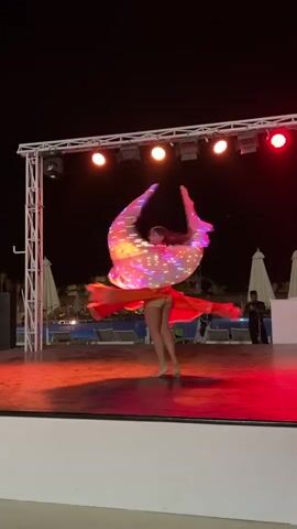 At night, a woman performs on a stage near a pool. She wears a sequined top, an orange skirt, and a see-through cape covered in white LED lights. The stage is lit with white and red stage lights. A steel light rig stands behind her. Behind the stage is a dark pool area with several white sun umbrellas and a water slide. A man in dark clothes stands to the right of the stage. She is dancing barefoot. She twirls and gestures with her arms to the music.