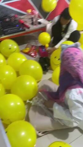 A group of children and adults are gathered in a room decorated with a red and white geometric pattern on one wall. Yellow balloons are scattered throughout the room, some with smiley faces drawn on them. One woman is holding a bunch of yellow balloons, while others are seated on the floor, interacting with the balloons. A young child in a pink dress is standing and looking at the balloons. The room appears to be a play area or party venue.
