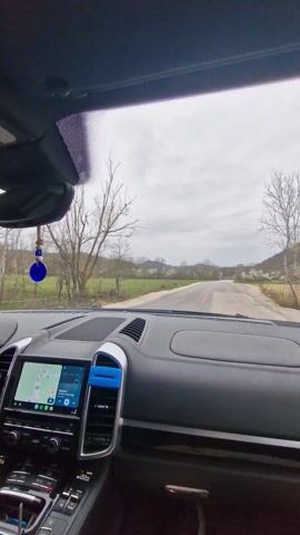Here's a description of the video:

The video shows the interior of a car, specifically the dashboard and front seats, while driving on a rural road. The car appears to be a Porsche Cayenne, given the dashboard design and steering wheel. The interior is mostly black with silver accents. There's a touchscreen display in the center console showing a navigation map and music information. A blue evil eye charm hangs from the rearview mirror.

The driver's hands are visible on the steering wheel. The view through the windshield shows a narrow, paved road winding through a landscape of green fields and scattered trees. The sky is overcast, suggesting a cloudy day. The camera angle varies slightly, sometimes focusing more on the interior of the car and other times more on the road ahead.