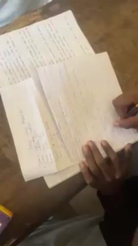 The video shows a close-up of someone writing on lined paper. They are using a black pen and appear to be taking notes on a wooden table covered with several pages of handwritten documents. The papers visible include a page titled "Single Stage Amplifier," detailing types of amplifiers: common base, common collector, and common emitter. Other pages contain handwritten text and equations. The person is wearing a black shirt and has a red bracelet on their wrist. The environment seems to be indoors, possibly a study or classroom setting.