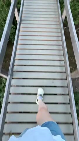 A person walks across a wooden bridge over a stream. They are wearing white clogs and denim shorts. The bridge is surrounded by green foliage. At the end of the bridge, a man stands on a gravel path, wearing a black t-shirt and shorts, holding a bag over his shoulder. The landscape features rolling green hills under a clear blue sky.