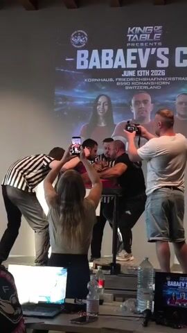 The video shows an arm wrestling match. Two men are seated at an arm wrestling table, facing each other, with their hands clasped in a grip. One man is wearing a black t-shirt and black pants, while the other is wearing a black and white striped referee shirt and grey pants. Spectators are gathered around the table, some of whom are recording the event with their phones and cameras. A large screen behind the arm wrestling table displays the "BABAEY'S CUP" logo and event details. The environment appears to be an indoor venue, possibly an arena or hall, with wooden beams visible on the ceiling. The lighting is dim, with spotlights focused on the arm wrestling table.