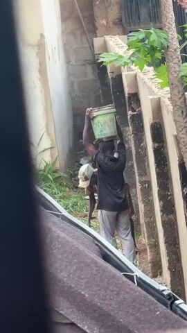 A man wearing a black t-shirt and gray pants carries a bucket on his head. He is walking away from the camera and towards a wall. Another person, partially obscured, is bent over in front of him. The environment appears to be an outdoor construction or work area with a brick wall and some vegetation. The camera is positioned above and behind the subjects, looking down.