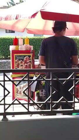 A street food vendor is seen preparing food under a red and white umbrella. The vendor is wearing a dark t-shirt and a cap. In front of the vendor is a food cart with condiments, fried food, and a menu. A motorcycle is parked behind the cart. In the background, there is a green hedge, a building, and trees. There are also other people visible in the background. The scene appears to be a sunny day.
