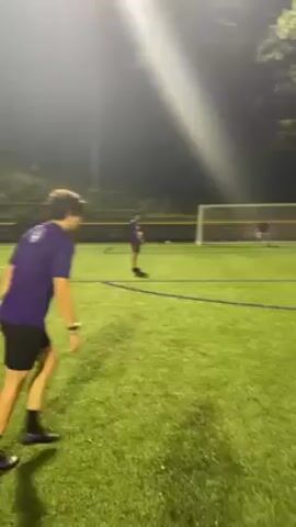 A soccer player in a purple shirt and black shorts kicks a soccer ball towards a goal, where a goalie stands ready. The kick occurs on a green artificial turf field at night, illuminated by bright stadium lights. Another player is seen in the background. The camera follows the ball's trajectory as it flies towards the goal.