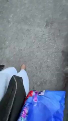 A handheld camera shows a person walking outdoors on what appears to be pavement or a sidewalk. The camera initially shows the person’s legs, clad in white pants, and their bare feet. They are carrying a black bag and a blue patterned tote bag. The camera then moves upward to reveal the person's face. They are wearing glasses, over-ear headphones, and a black jacket over a grey hooded sweatshirt. The background shows grass and trees, suggesting a park or green space. The person seems to be recording themselves while walking.