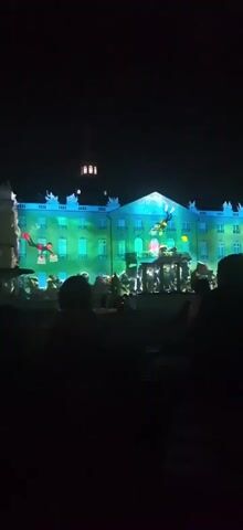 The video captures a nighttime outdoor performance in front of a large building adorned with bright green and blue lights. Multiple figures are projected onto the building, appearing to be diving or flying through the air. In the foreground, silhouettes of people are visible, suggesting an audience watching the spectacle. The atmosphere is dark, with the building and projected figures being the primary sources of illumination.