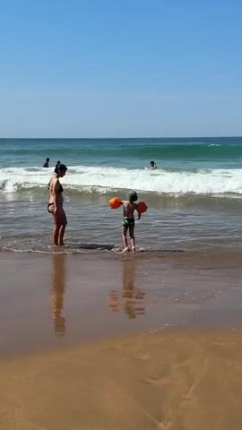 The video captures a sunny day at Zarautz beach in Spain. The scene is set on a sandy beach with gentle waves rolling in from a clear turquoise ocean. The sky is a vibrant blue, suggesting fair weather.

In the foreground, a woman in a black bikini stands in the shallow water, facing away from the camera, with her reflection shimmering on the wet sand. Beside her, a child wearing a green hat and orange arm floaties stands with their back to the camera, also facing the ocean. The child appears to be interacting with the incoming waves, perhaps about to splash or play.

Further out in the ocean, several other people are visible enjoying the water. One person is partially submerged, and another is swimming or wading further out. The waves are moderately sized, creating white foam as they break near the shore. The overall atmosphere seems relaxed and enjoyable, indicative of a typical beach day. The lighting suggests it is daytime, likely midday or early afternoon due to the bright sun. There is no visible text in the media file.