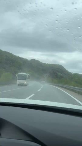 The video shows a car driving on a road, with rain falling on the windshield. Windshield wipers can be seen clearing the rain. The road is wet and the sky is overcast. There are mountains on either side of the road. Other cars can be seen driving on the road as well.