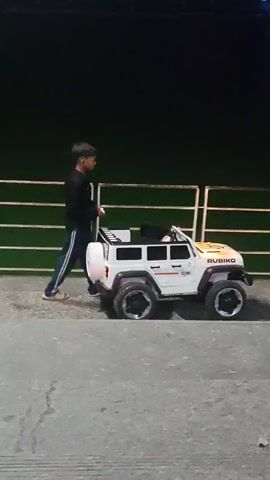 A young boy is standing next to a white toy car that appears to be a Jeep. He is holding something in his hands and seems to be interacting with the toy car. The car has a spare tire mounted on the back and the word "RUBIKO" is visible on the side. The scene takes place outdoors at night, with a dark green fence or wall in the background and a concrete ground. There are some lights visible in the distance in the upper right portion of the frame, possibly indicating a cityscape or a well-lit area. The boy is wearing a black long-sleeved shirt and blue jeans.