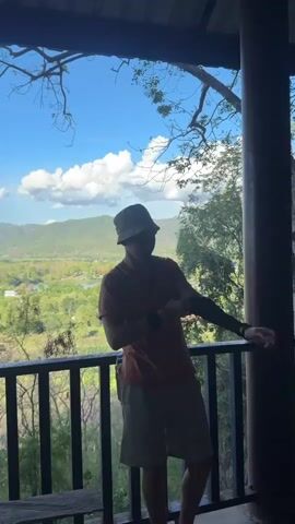 The video shows a man standing on a balcony, facing away from the camera, and looking out at a lush green valley dotted with trees and a lake. He is wearing a hat, a t-shirt, shorts, and a black arm sleeve. The balcony has a dark wooden railing. The sky is blue with fluffy white clouds. The camera zooms in on the view of the valley.