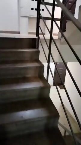 The video shows a first-person perspective of someone ascending a concrete staircase with a metal railing on the right. The staircase appears to be indoors, likely in a building's common area such as a hallway or stairwell. The walls are painted white, and the floor is tiled with square, beige tiles. A white electrical panel with several compartments and a yellow warning triangle is visible on the wall to the right of the staircase. A dark brown door is also visible to the right of the electrical panel. The lighting is artificial and casts shadows, suggesting an indoor environment.