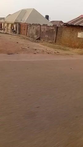 Here's a description of the video:
The video is shot from a moving vehicle traveling on an asphalt road. The environment appears to be a residential or commercial area with buildings lining the sides of the road.
Several details are visible:
*   Buildings: There are various buildings including a structure with a water tank on top, small shops or stalls with goods displayed, and houses.
*   People: People are seen walking on the roadside. One man is carrying items on his head.
*   Objects: A pile of trash/rubbish is noticeable near the roadside.
*   Environment: The scene looks somewhat dusty. The sky is overcast.