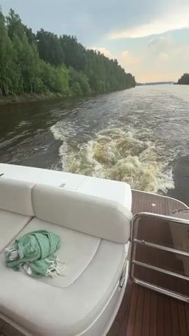 The video shows a view from the stern of a white boat moving through a wide, muddy river. Trees line the left bank of the river, and the right bank is less densely wooded. The sky is overcast with some lighter areas suggesting the sun may be breaking through. A green scarf is draped over a white cushion on the boat's seating area. The boat is creating a large wake behind it, with foamy, churning water. The overall impression is of a leisurely boat trip on a cloudy day.