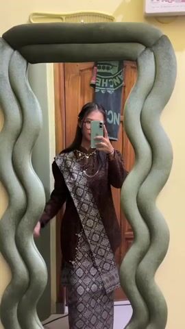 A young woman with long dark hair and glasses stands in front of a mirror with a wavy, plush green frame, taking a selfie with her light green smartphone. She is wearing a long-sleeved dark brown top, a patterned long skirt or sarong in shades of brown and silver, and a matching patterned sash draped over her left shoulder and across her body. A necklace is visible around her neck. Her expression is a slight smile. The background behind her in the mirror's reflection shows a yellow wall and a wooden door with a dark green cloth hanging on it.