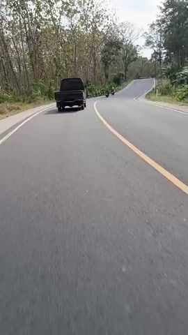 A black pickup truck is driving on a paved road. The road is curved and uphill, with trees and foliage on either side. There are two motorcycles in front of the truck. The sky is overcast.