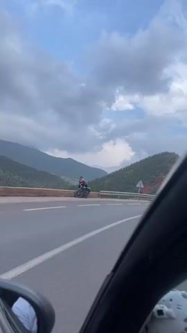The video captures a scenic drive through a mountainous region in Taljarft, Morocco, from the perspective of a moving vehicle.

At the beginning (0:00-0:01), a person wearing a red and black jacket and a helmet is seen parked on a dark motorcycle on the side of a two-lane road. A metal guardrail separates the road from a steep drop-off. In the background, lush green mountains rise under a cloudy sky with patches of blue. Part of the car's side mirror is visible in the bottom left, indicating the recording location.

As the vehicle moves forward (0:01-0:02), the motorcyclist disappears, and a triangular warning sign with a black arrow comes into view behind the guardrail. The landscape continues to feature rolling, green-clad mountains with some exposed reddish-brown earth.

The view then pans across the mountainous terrain (0:02-0:03), showcasing deep valleys and diverse vegetation patterns on the slopes.

Finally (0:03-0:04), a traditional village is revealed nestled within a valley among the mountains. The buildings appear to be constructed from reddish-brown earth, and a prominent mosque structure with a minaret is visible. Another road sign, a white rectangular one with a black arrow indicating a sharp left curve, is seen behind the guardrail. The sky remains mostly cloudy throughout the video, which appears to be recorded during daytime.