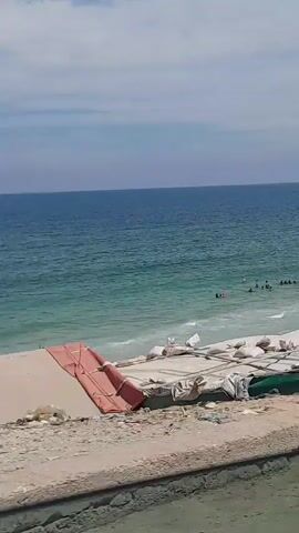 The video shows a beach on a bright day. The sky is blue with some clouds. The ocean is a mix of light blue and turquoise and has small waves.  A group of people, mostly silhouettes, are in the water. They appear to be swimming and playing. A sandy beach stretches out towards the water. In the foreground there is a structure with a tarp-like covering. As the camera moves, a white building structure comes into view, partially obscuring the view of the beach. A lone figure walks along the beach.