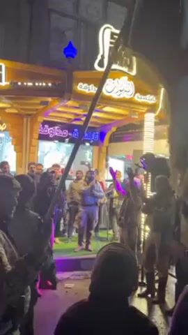 A group of people is gathered on a street at night. A person in the foreground holds a flag with a red star on it.  Behind them, other people are standing and watching a group of men on a slightly raised platform. Some of the men on the platform are wearing military-style clothing. One man holds a microphone and appears to be speaking or singing.  A building with bright lights and Arabic writing on it is in the background. The crowd is illuminated by streetlights and the lights from the building. Some people in the crowd are recording the event with their phones.