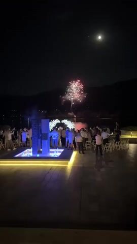 The media file captures an outdoor event at night, likely a wedding ceremony or celebration, in Razliv, Bulgaria. The scene is illuminated by a combination of artificial lighting and fireworks. A group of people, dressed in formal attire, are gathered. In the foreground, there is a modern fountain with purple and blue lighting. Behind the fountain, a white floral arch stands, serving as a focal point for the ceremony.

The main activity is a fireworks display occurring in the background, with multiple bursts of light and smoke visible against the dark night sky. The moon is also visible, high in the sky. The setting appears to be a paved outdoor area, possibly a terrace or plaza, with rows of white chairs set up, suggesting an audience or guests. In the distance, mountains and a body of water can be seen, hinting at a scenic location.

The time of day is clearly night, with a dark sky and artificial lighting playing a significant role in illuminating the scene. The weather appears clear, as indicated by the visible moon and the lack of precipitation. The overall mood conveyed is festive and celebratory, enhanced by the fireworks and the gathering of people. There is no visible text in the media file.
