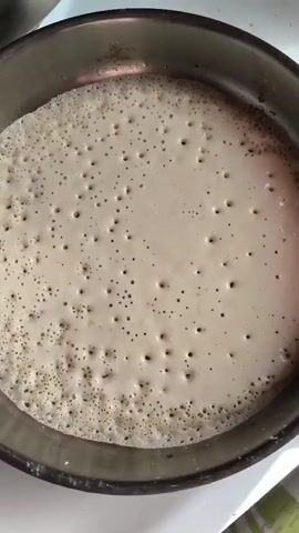 The video shows a close-up, overhead view of a batter cooking in a pan. The batter is a light beige color and is covered in small, evenly distributed bubbles, indicating that it is being heated and is likely a type of pancake, crepe, or dosa. The pan appears to be made of metal and has a slightly worn, perhaps non-stick, surface. The lighting suggests an indoor environment, possibly a kitchen. The camera is held steadily, focusing entirely on the cooking batter.