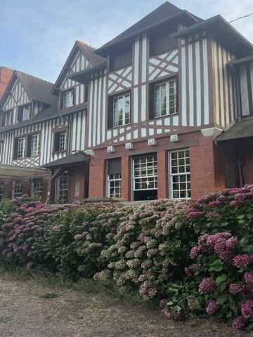 This video shows a half-timbered building with red brick accents, fronted by a dense hedge of pink and purple hydrangeas. The building has multiple stories and dormer windows. To the left of the building, a white tent structure is partially visible, and further left, a person in a white shirt and pants stands on a gravel path facing away from the camera. Behind the building, a yellow and blue striped circus tent is visible, with a French flag flying nearby. The scene transitions to show a vintage tan truck with a green tarp cover parked in a grassy area. The truck has "CIRCUS" written on its side. Other circus-related equipment, such as tables and chairs, are visible in the background. The environment is wooded, with tall trees framing the scene. The sky is clear and blue.