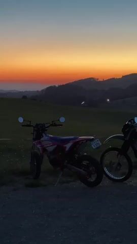 The video shows two dirt bikes parked on a gravel path next to a green grassy field. The bikes are silhouetted against a vibrant sunset sky with hues of orange, yellow, and blue. Rolling hills are visible in the background, and a dark line of trees is on the left side of the frame. The video transitions to a blurry shot of a road leading away from the sunset, with the trees and hills becoming indistinct.