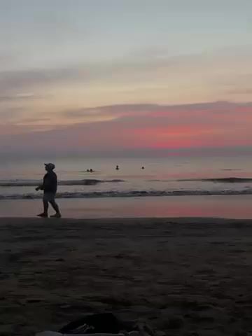 A man is walking on a beach at sunset. He is wearing a hat, a dark shirt, and light shorts. He is carrying something in his hand. There are a few other people in the water. The sky is a mix of pink, orange, and gray. There are some personal belongings lying in the sand.