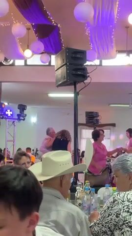 The video shows a reception hall full of people celebrating. Some guests are seated at tables, while others are dancing to music. There is a large speaker on a stand, and the room is decorated with white and purple fabric, string lights, and paper lanterns. The architecture features brick columns and windows along the top of the walls.
