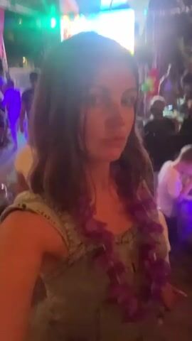 A woman with brown hair and a purple lei around her neck is filming herself with a cell phone in selfie mode. She is at an outdoor party or event at night. In the background, there are lights, a stage with a screen, decorations, and other people dancing and sitting at tables. The woman is wearing a denim dress. She appears to be talking to the camera.