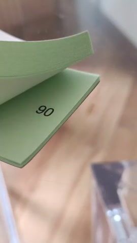 The video shows a close-up of a pad of small, light green paper sheets on a shiny, reflective surface. The sheets are numbered sequentially. Someone flips through the sheets, revealing numbers from 73 to 01. The background is blurred, but appears to be a wooden surface with a small, clear plastic container on it.