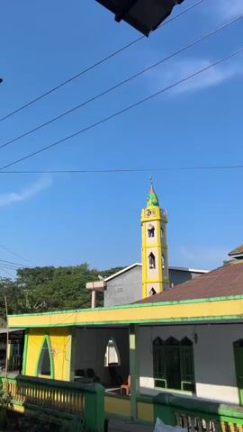 The video begins with a view of a mosque with a tall, yellow and green minaret against a bright blue sky. The camera pans slightly to the right, revealing the main structure of the mosque with a brown, corrugated metal roof and a dome. In the foreground, there is a low wall or railing painted green and yellow, with white walls and green-framed windows and doors belonging to a building adjacent to the mosque. The camera continues to pan right, showing more of the building's facade and lush green trees and vegetation on a hillside in the background. Finally, the camera pans further to reveal a utility pole with overhead wires and more trees. The overall scene depicts a mosque and its surroundings on a sunny day, with clear blue skies and abundant greenery. There are no people or specific actions visible in the video.