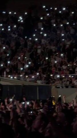 The video captures a concert scene. The foreground shows a crowd of people, many holding up their phones with their flashlights on, creating a sea of small lights. The middle ground reveals a stage area with some equipment and figures, possibly crew members. In the background, the audience continues, with more lights from phones scattered throughout the seating areas. The environment appears to be a large venue, likely an arena or stadium, with tiered seating. The lighting is predominantly dark, illuminated by the phone lights and some stage lighting, suggesting it is nighttime or an indoor event. The overall atmosphere conveyed is one of excitement and engagement from the concertgoers.