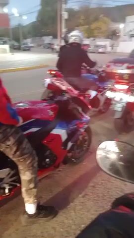 The video shows a group of motorcyclists stopped at a traffic intersection. There are at least three motorcycles visible, primarily red with white and blue accents. One has a GIVI storage box mounted on the back. Riders wear helmets and casual clothing. The camera view is from the perspective of a motorcyclist. The environment is an urban street with buildings, utility poles, and other vehicles in the background. Pedestrians cross the street. The sky is overcast. The general scene suggests a casual gathering or ride of motorcyclists.
